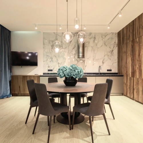 „Modern eingerichteter Essbereich mit rundem Holztisch, sechs dunkel gepolsterten Stühlen, dekorativem Blumenarrangement und offener Küchenzeile mit Marmor-Rückwand und Hängelampen – elegantes Wohnambiente mit hochwertigem Interieur.