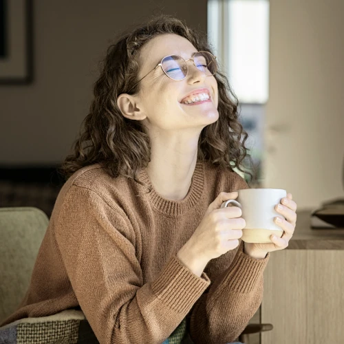 Zufriedene Frau mit Brille und braunem Pullover hält eine Tasse in den Händen und lächelt entspannt – Symbolbild für Wohlbefinden, Kundenzufriedenheit oder positive Erfahrungen.