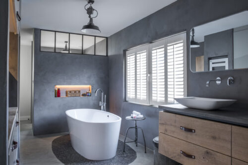 Modernes Badezimmer mit freistehender Badewanne, großem Spiegel, Holzschrank und Fenster mit weißen Jalousien.