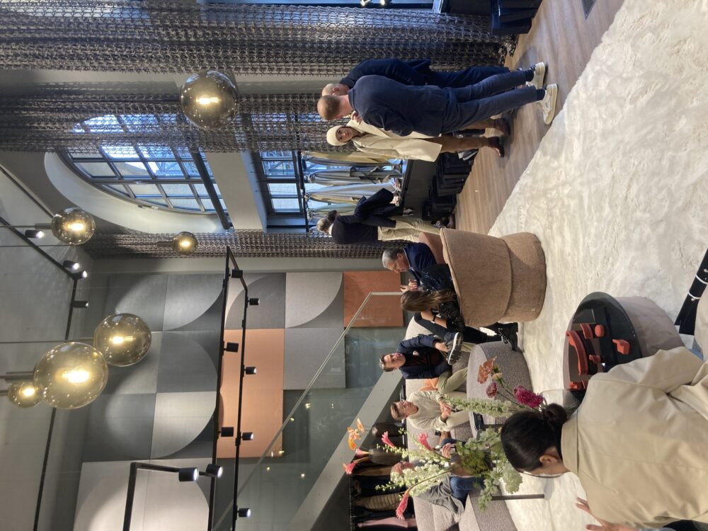 Moderne Lounge mit großen Fenstern, mehreren Personen im Gespräch und stilvoller Beleuchtung.