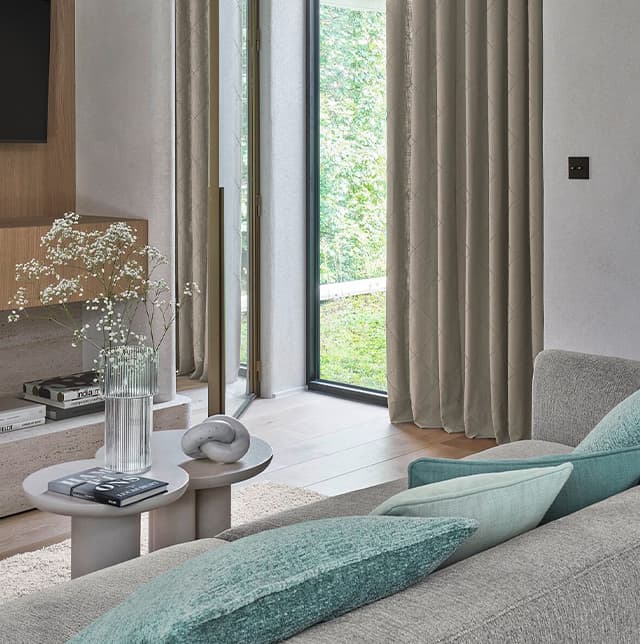 Moderner Wohnraum mit grauem Sofa, türkisfarbenen Kissen, kleinen Tischen und großen Fenstern mit Vorhängen.